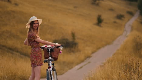 Back-kind-of-Happy-blonde-girl-in-dress-and-hat-turns-around-and-smiling-cheerfully-looks-at-the-camera-and-flirts-strolling-around-the-field-in-summer-with-bike-and-flowers.